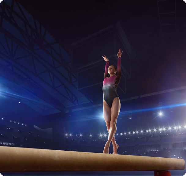 A gymnast balances gracefully on a beam in a large, brightly lit arena. She wears a sparkling leotard, with spotlights highlighting her poised form.