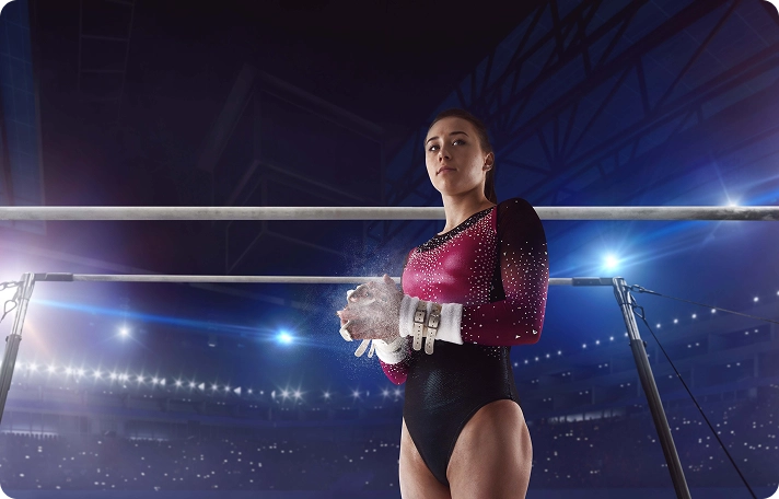 A gymnast in a red and black leotard stands confidently before uneven bars, chalking her hands. The stadium is dimly lit, creating a dramatic atmosphere.