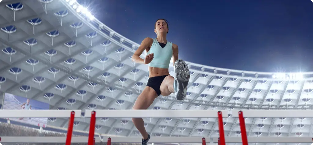 Athlete mid-air leaping over hurdle in a stadium, under bright lights. She wears a light athletic outfit, showing determination and focus.