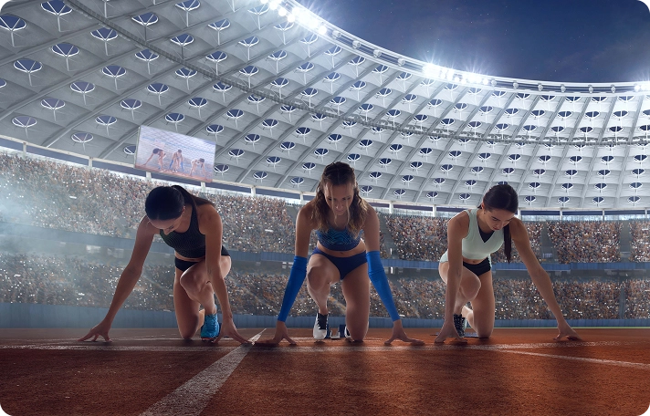 Three female athletes in starting positions on a track, under stadium lights, with a large crowd in the stands. The scene is tense and anticipatory.